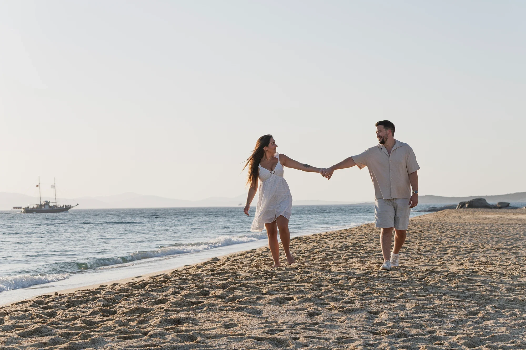 Couple walking hand in hand on a Naxos beach at golden hour with a sailboat in the distance
