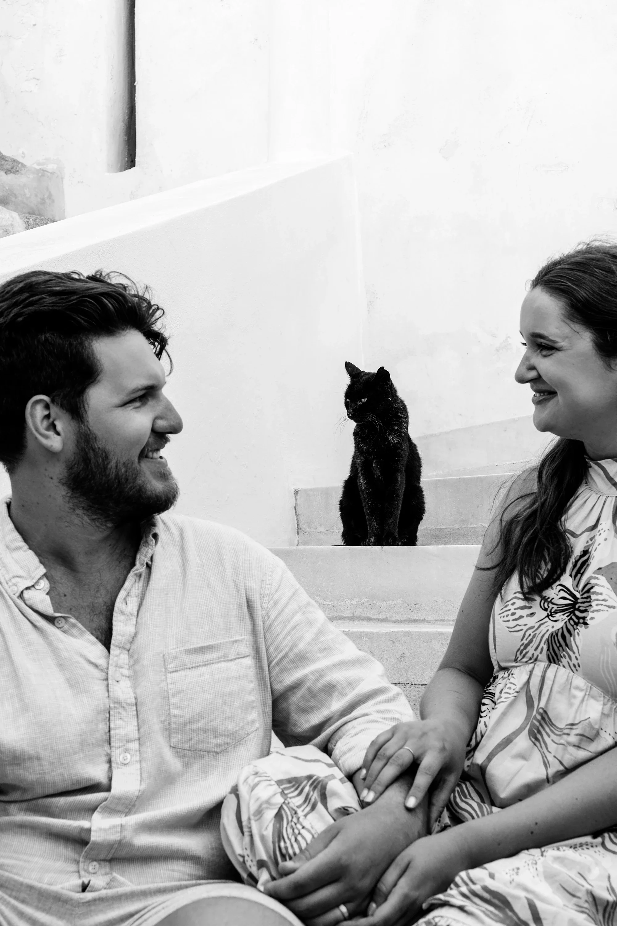 Black and white portrait of a couple smiling at each other with a cat on the stairs in Naxos