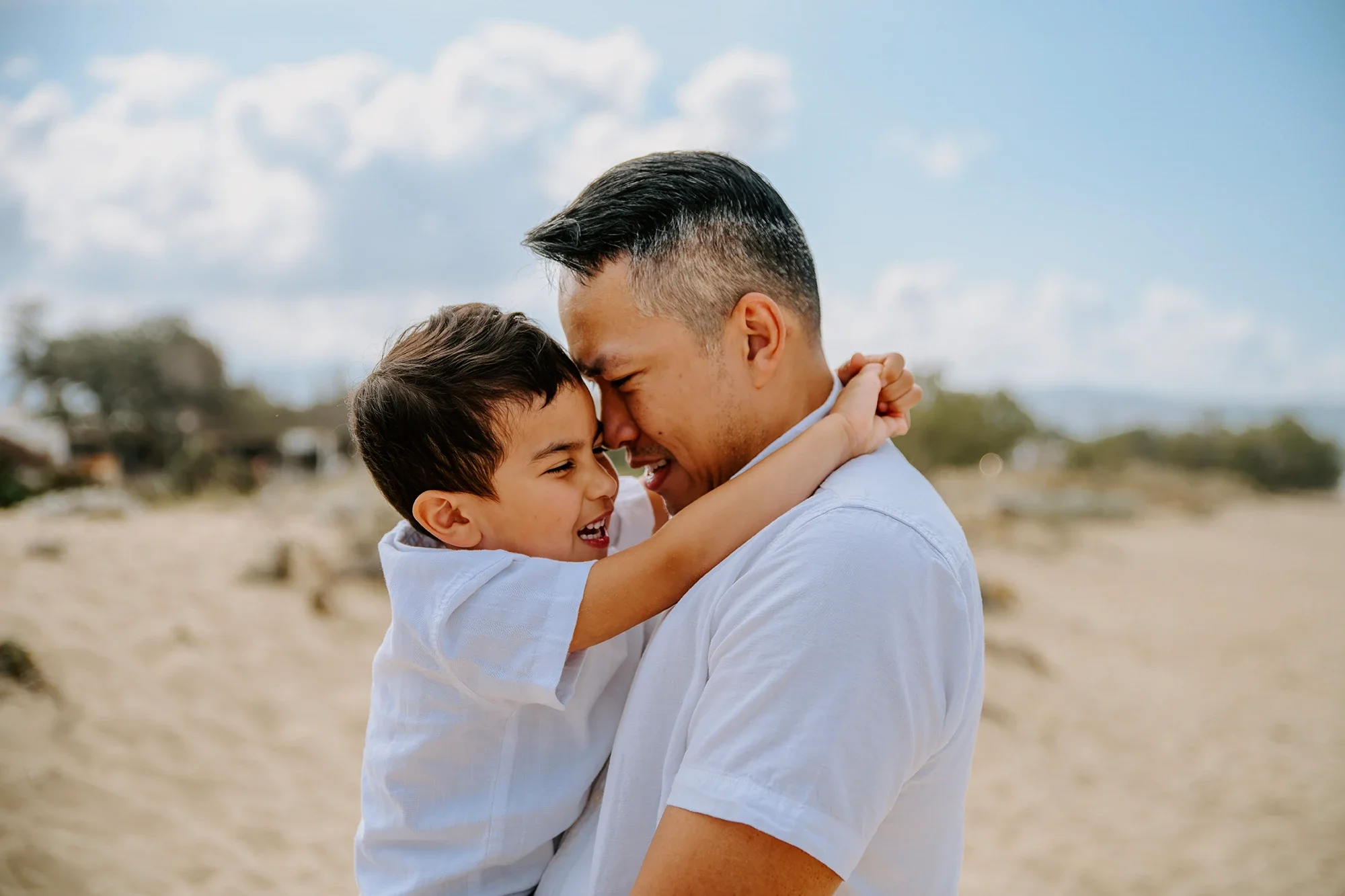 Family photography in Naxos: a father and young son share a laughing embrace on a sandy beach, with dunes behind them.