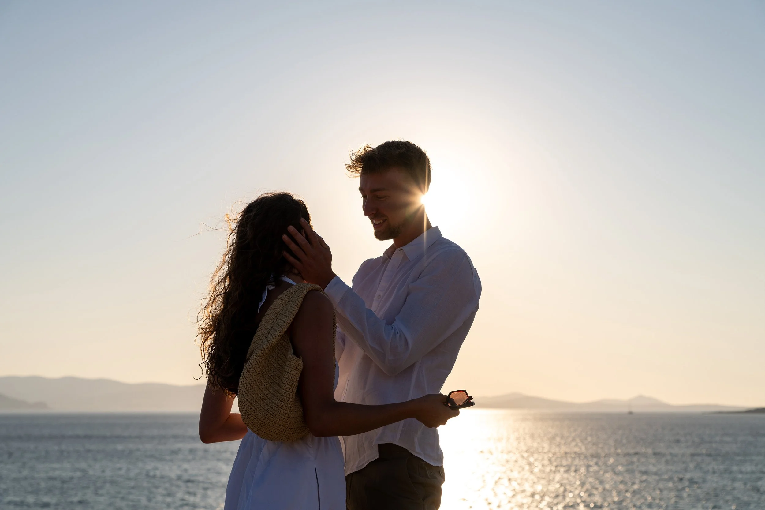A man gently touching a woman's face during sunset by a body of water, with mountains in the background.