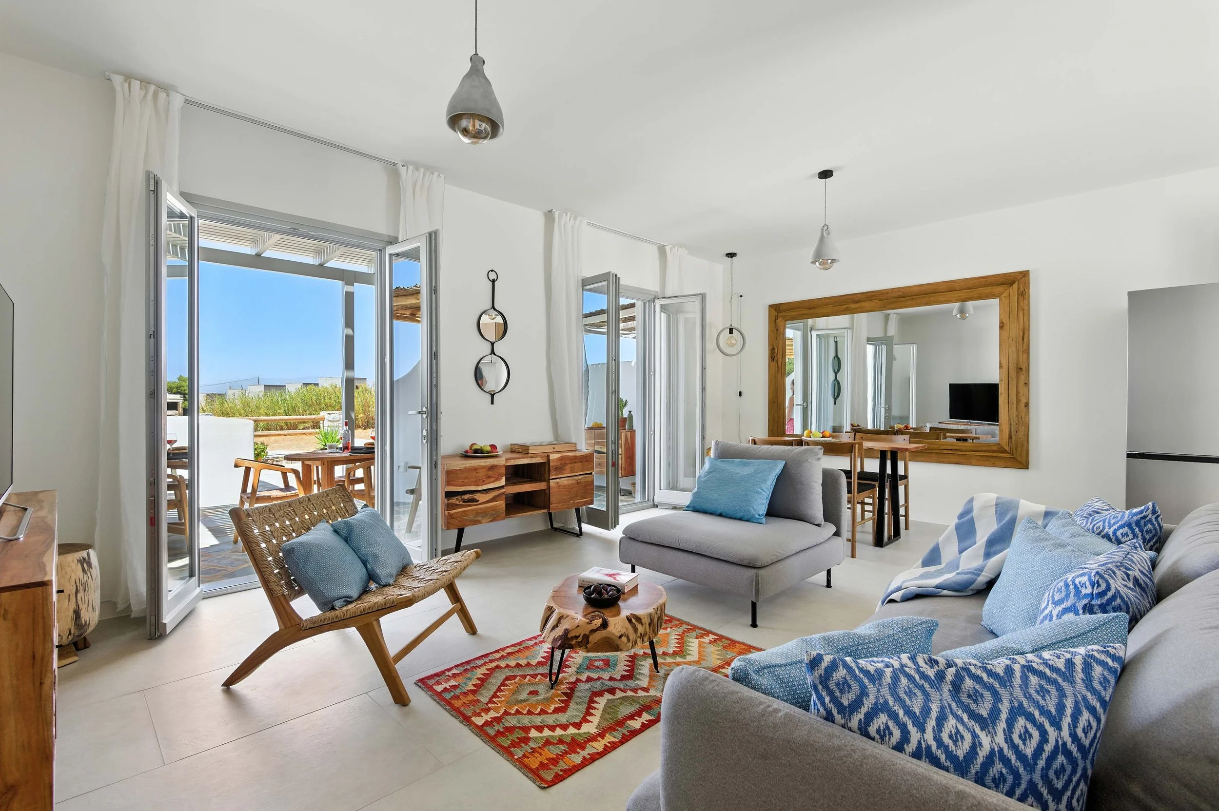 Bright and airy living room in Kastraki, Naxos with Cycladic-style decor, wooden accents, and large glass doors opening to a sunlit terrace – ideal for showcasing vacation rentals, luxury villas, and real estate listings.