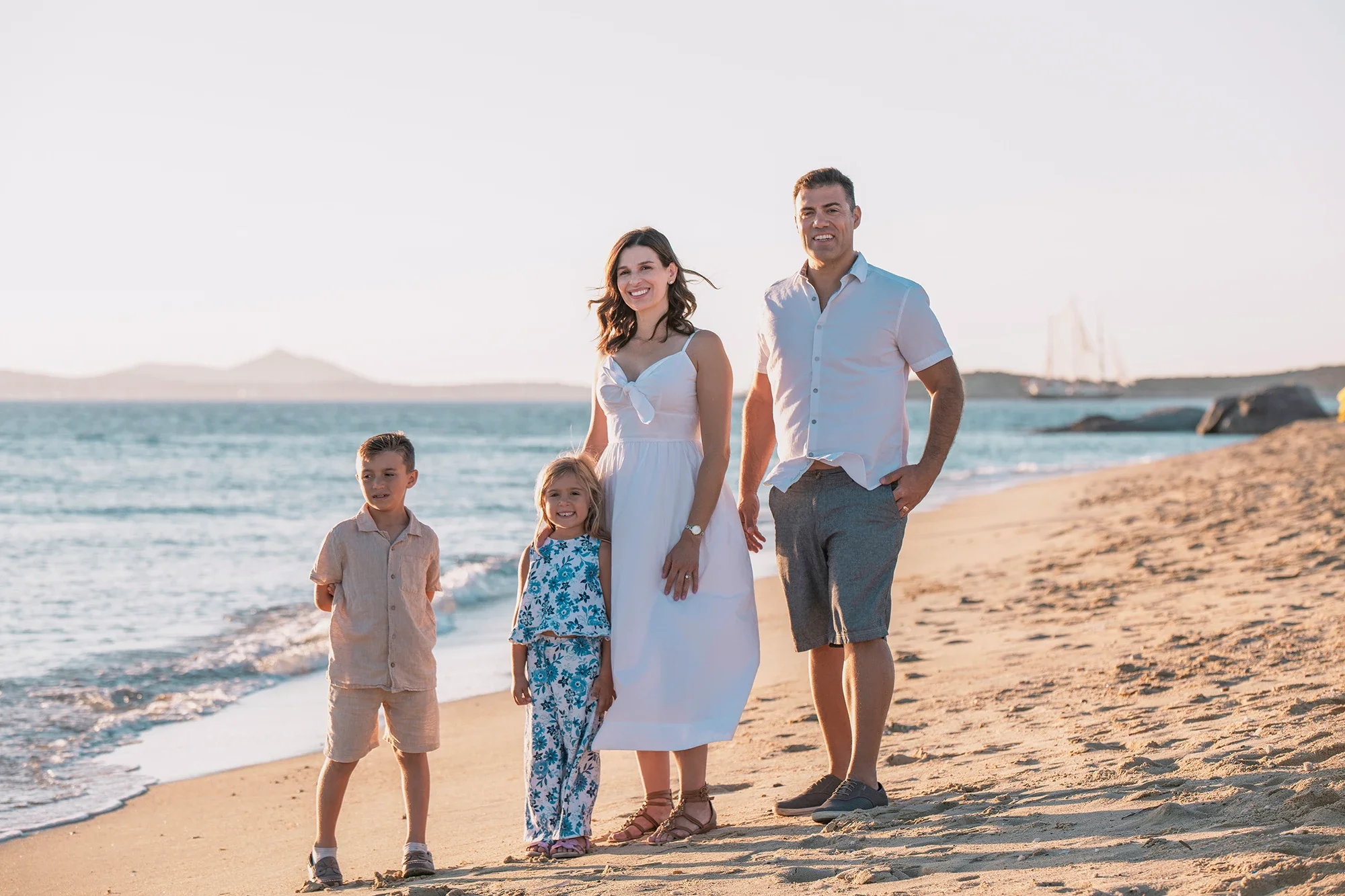 Family of four on a Naxos beach during golden hour with sea and sailboat in the background