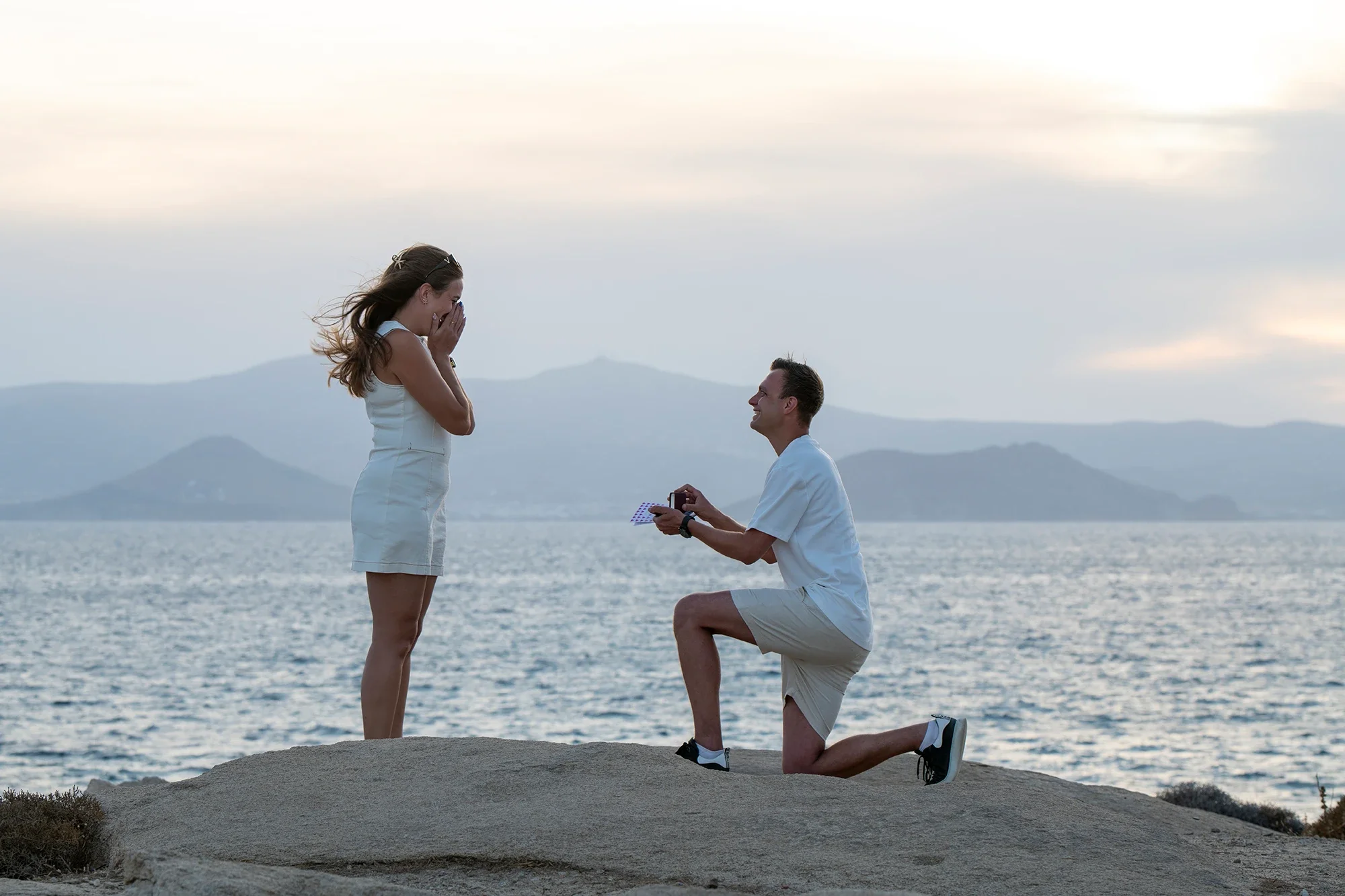 Woman covering her face in surprise during a proposal by the sea in Naxos