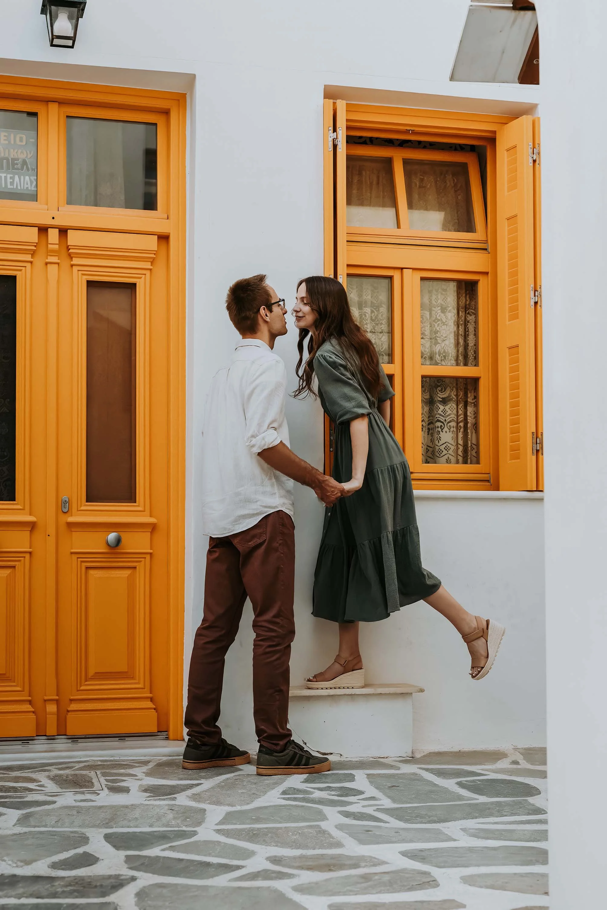 Young couple holding hands and smiling in front of a traditional white house with vibrant orange shutters in Naxos Old Town – romantic honeymoon photoshoot by Naxos Photographer, 2025.