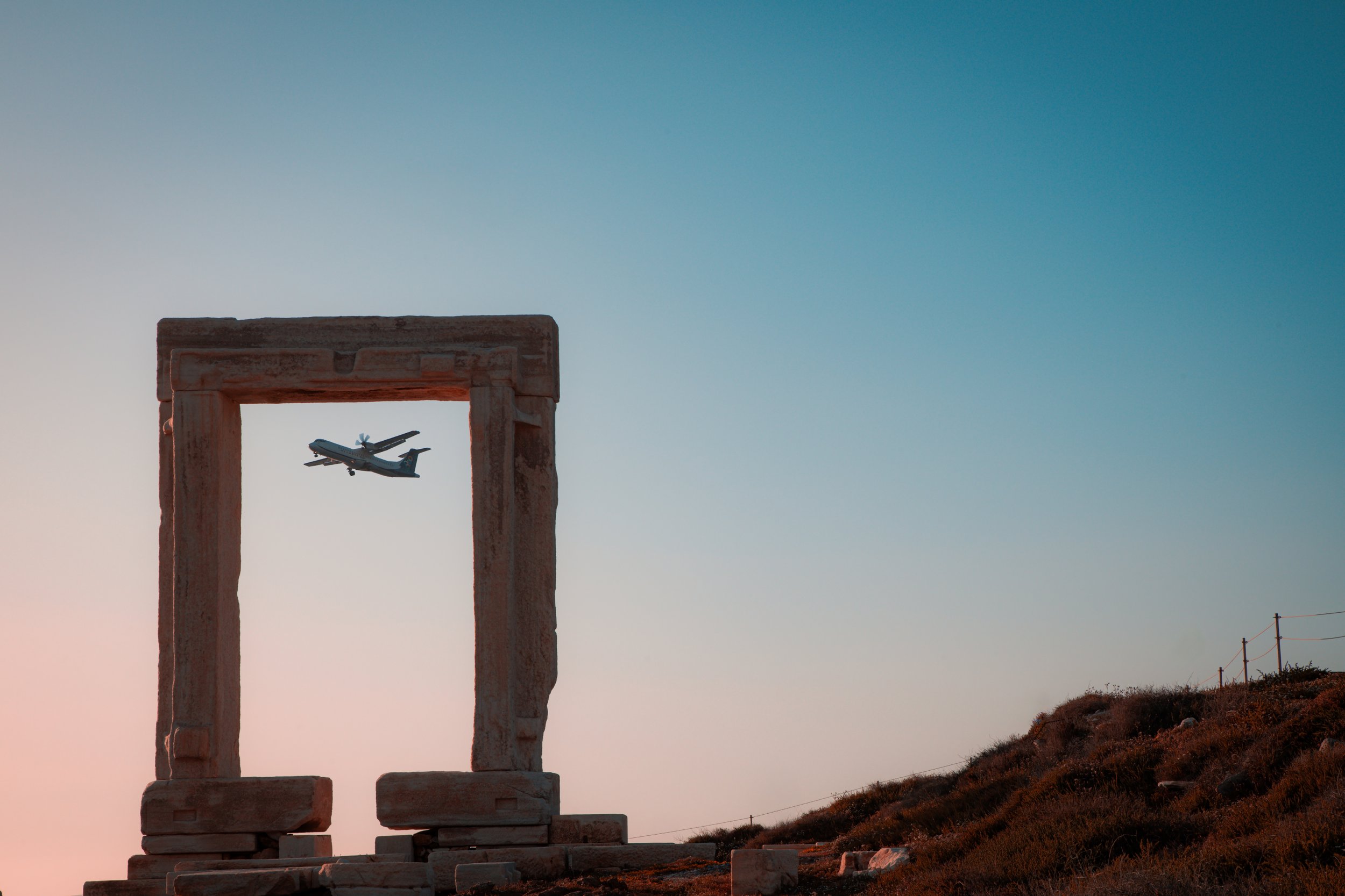 Airplane flying through the Portara of Naxos at sunset, ancient Greek marble gate with clear sky in the background, iconic travel and tourism landmark in Naxos, Greece. Picture taken by Spyros Plakidas for Naxos Photographer in April, 2025.