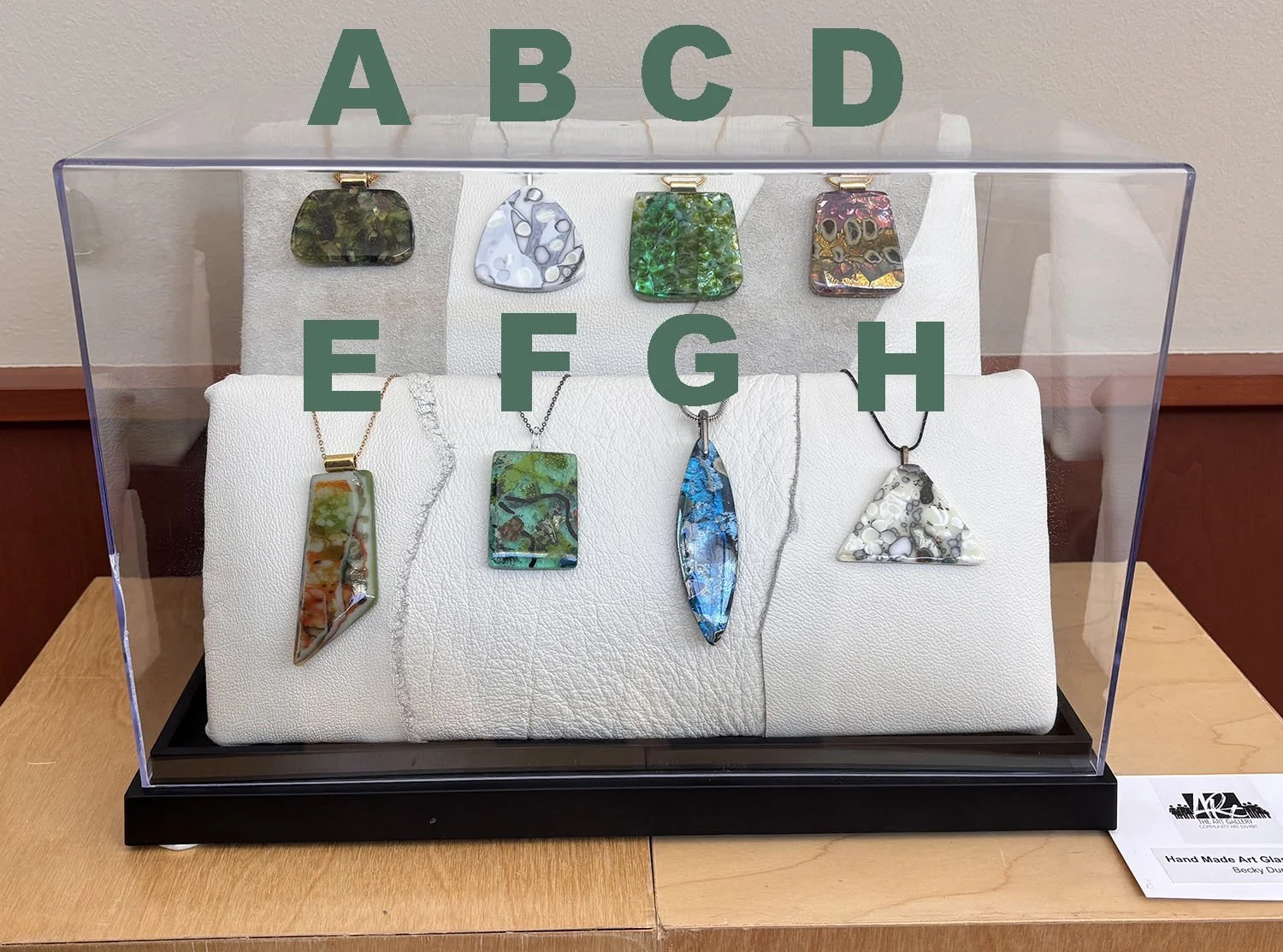 Hand Made Art Glass Jewelry,
Created by Becky Dunn.
From Left to Right, Top to Bottom of Display case: 
A.	Fused Art Glass Pendant with gold plated 16” mesh chain, $58.
B.	Fused Art Glass Pendant using glass which is reacting to the glass it touches 