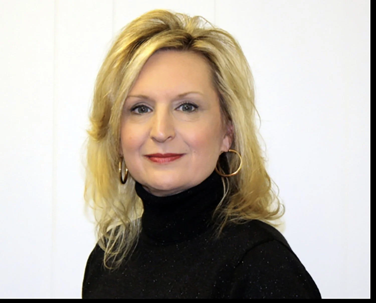 A woman with blonde, shoulder-length hair, wearing a black turtleneck sweater with subtle sparkles, gold hoop earrings, and red lipstick, standing in front of a plain white wall.