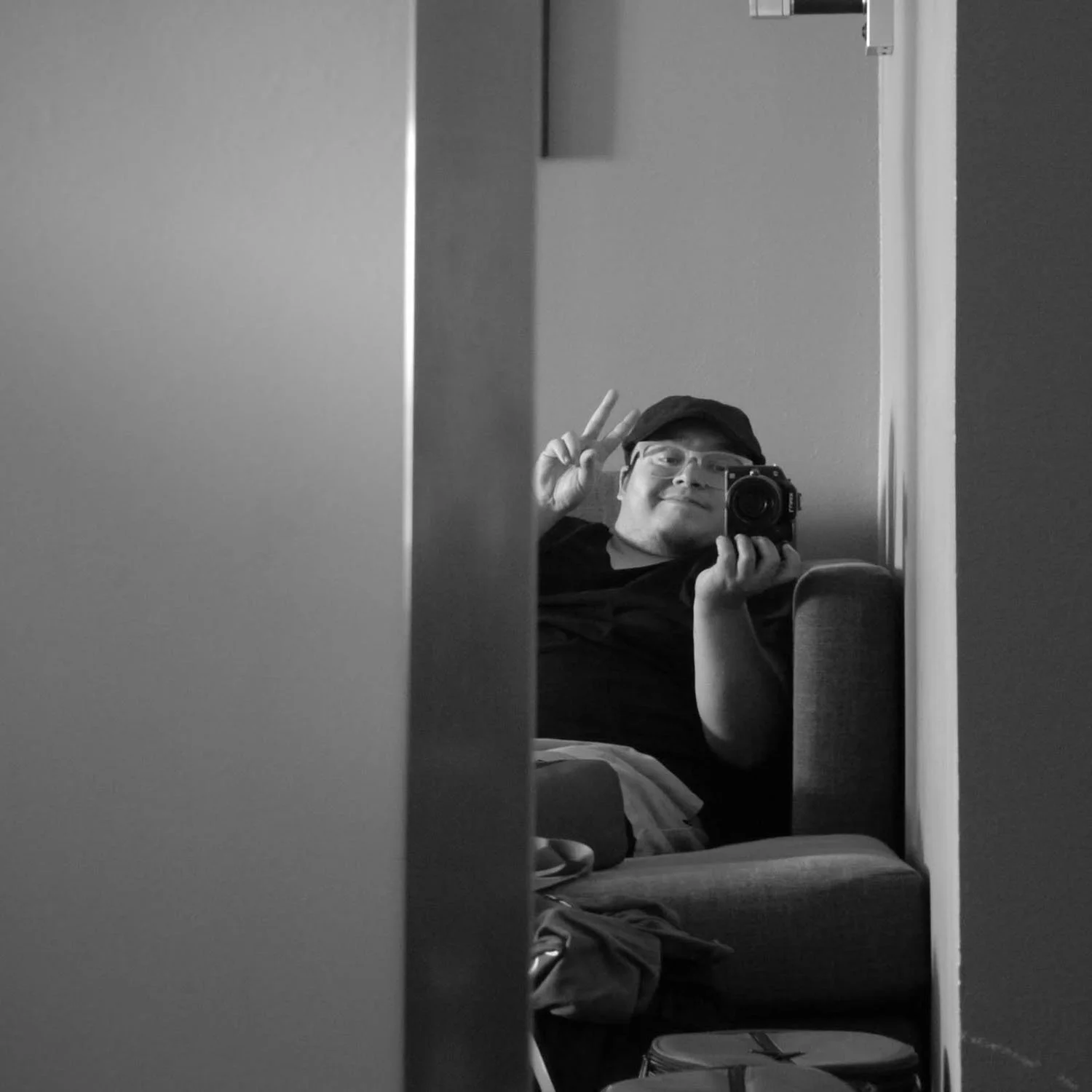 Person taking a selfie in a mirror, making a peace sign with one hand, wearing glasses and a cap, in black and white