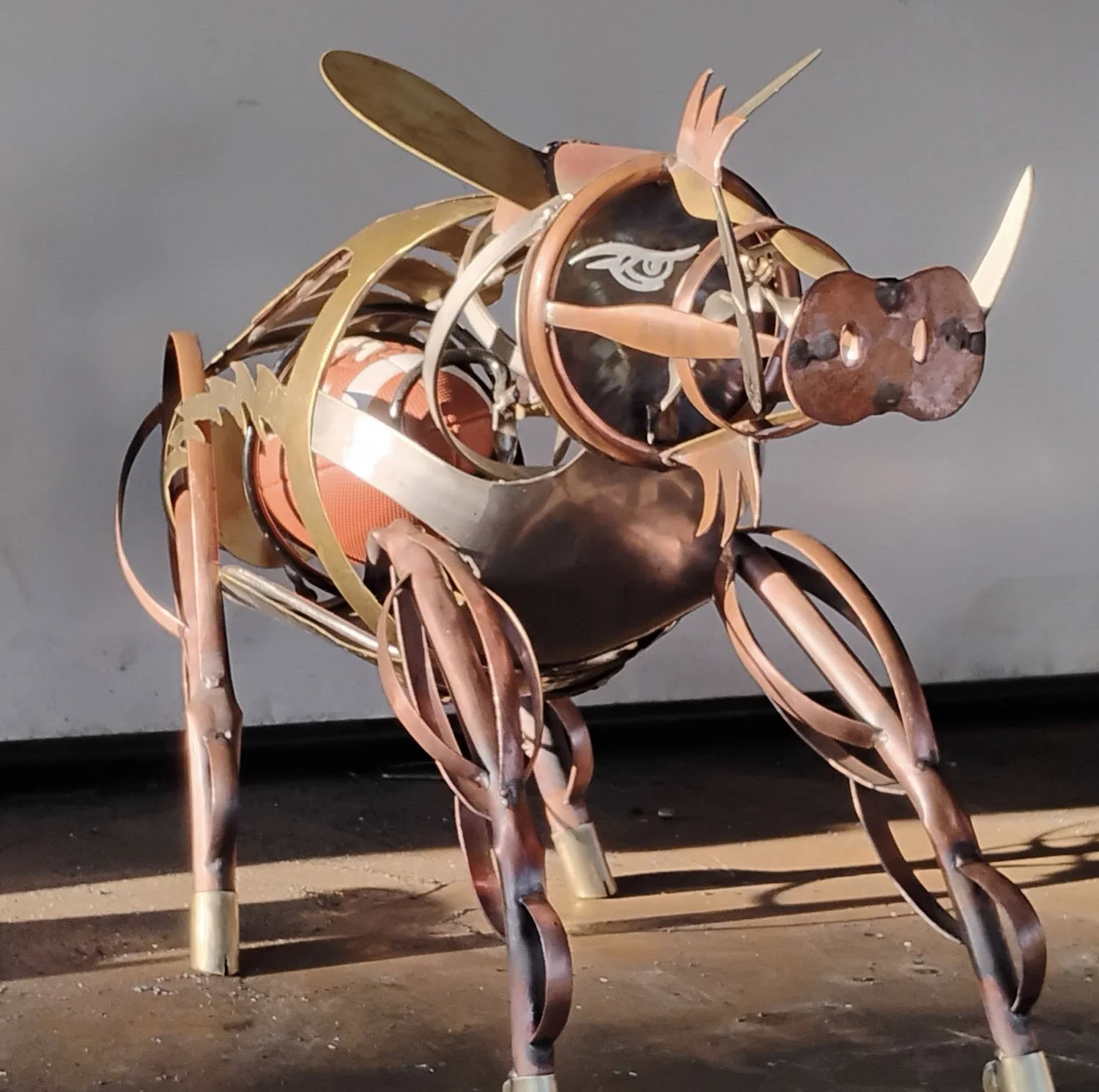 Metal sculpture of a pig with a spherical body, four legs, a snout, ears, and a tail, made from various metal pieces, set against a plain background.