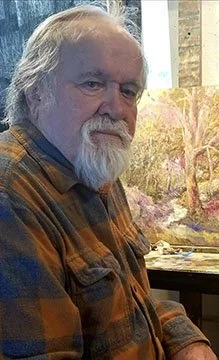 An older man with white hair and a beard, wearing a plaid shirt, sitting in front of a painting of a landscape with trees and a pink sky.