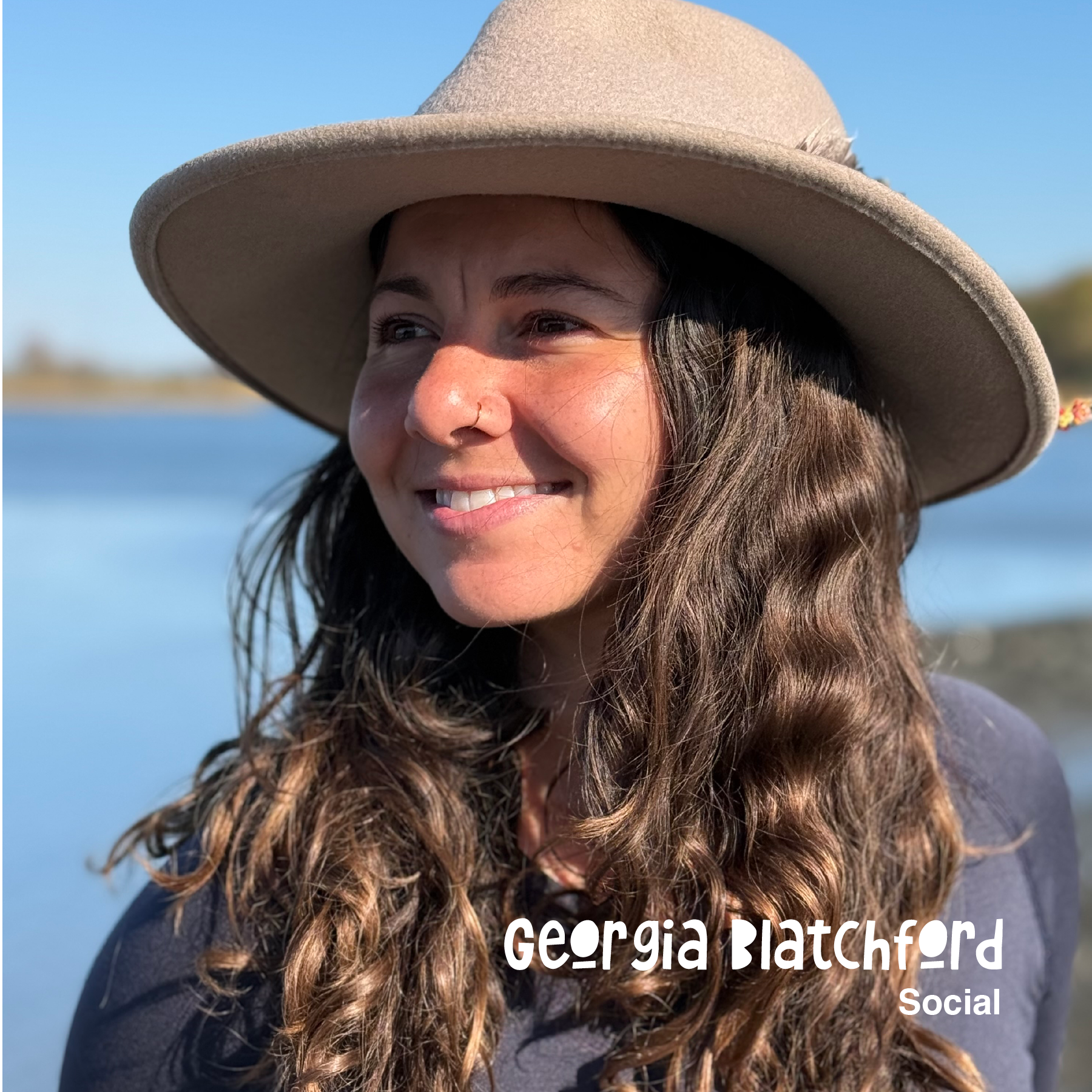 Close-up of a woman with long wavy brown hair wearing a beige wide-brimmed hat and a dark top, outdoors near a body of water under a clear blue sky, smiling.