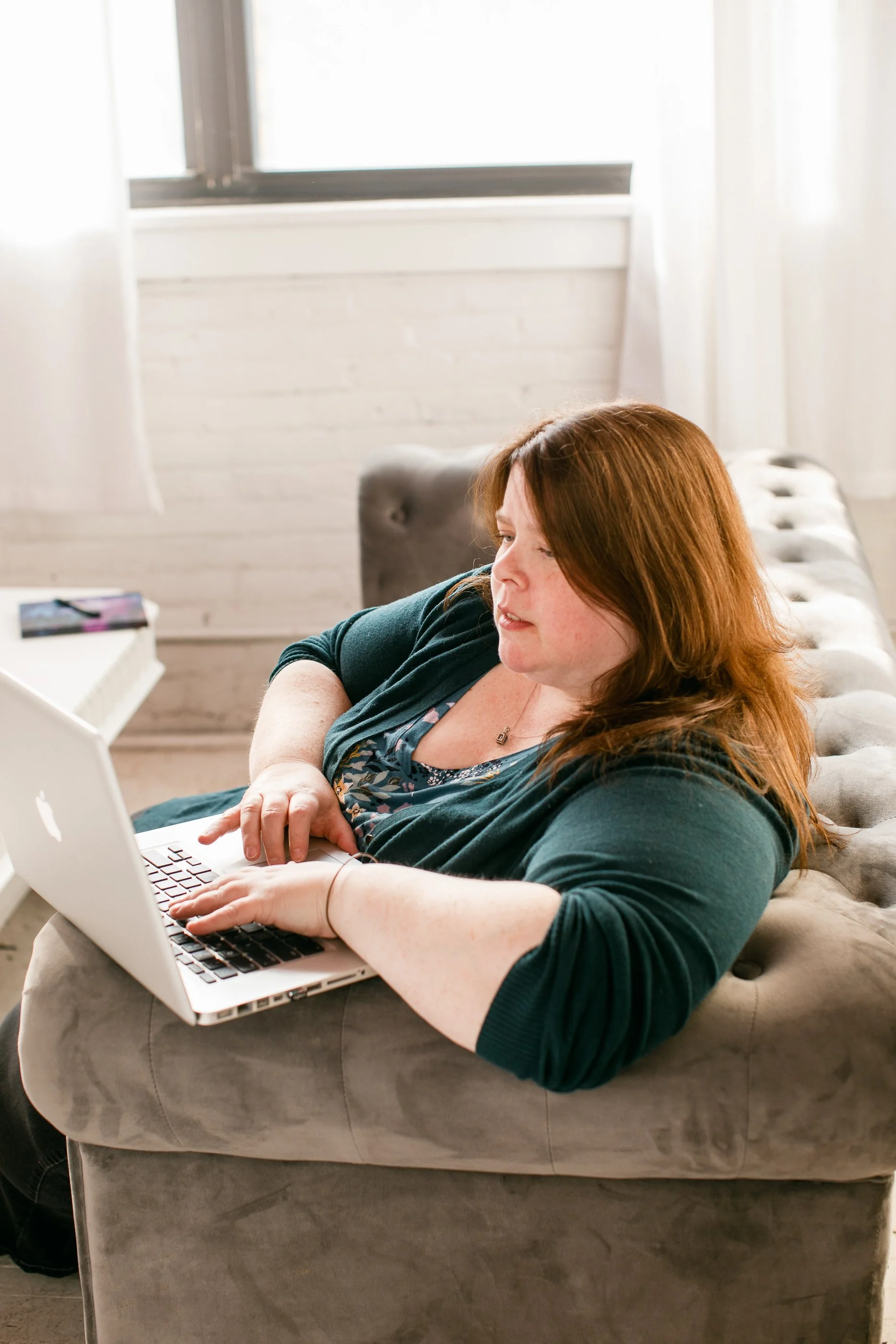 A photo of Melanie sitting on a gray couch using her laptop