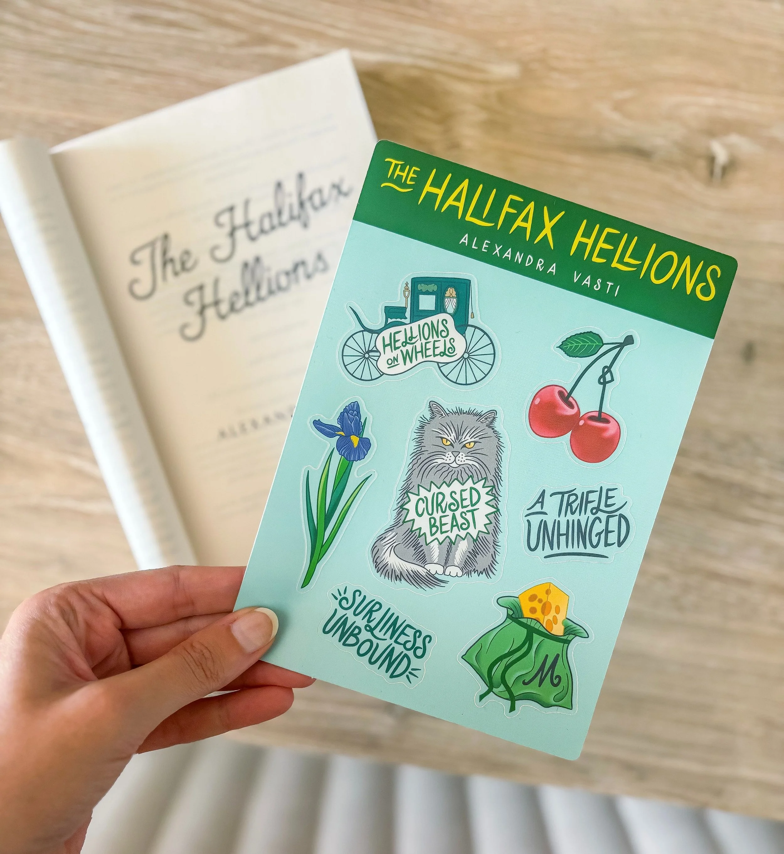 Hand holding a sheet of colorful stickers featuring characters and themes from 'The Halfax Hellions,' including a wagon with the team name, cherries, an iris flower, a snarling wolf labeled 'Cursed Beast,' text phrases, and a cheese wedge, with a blurred open book in the background.