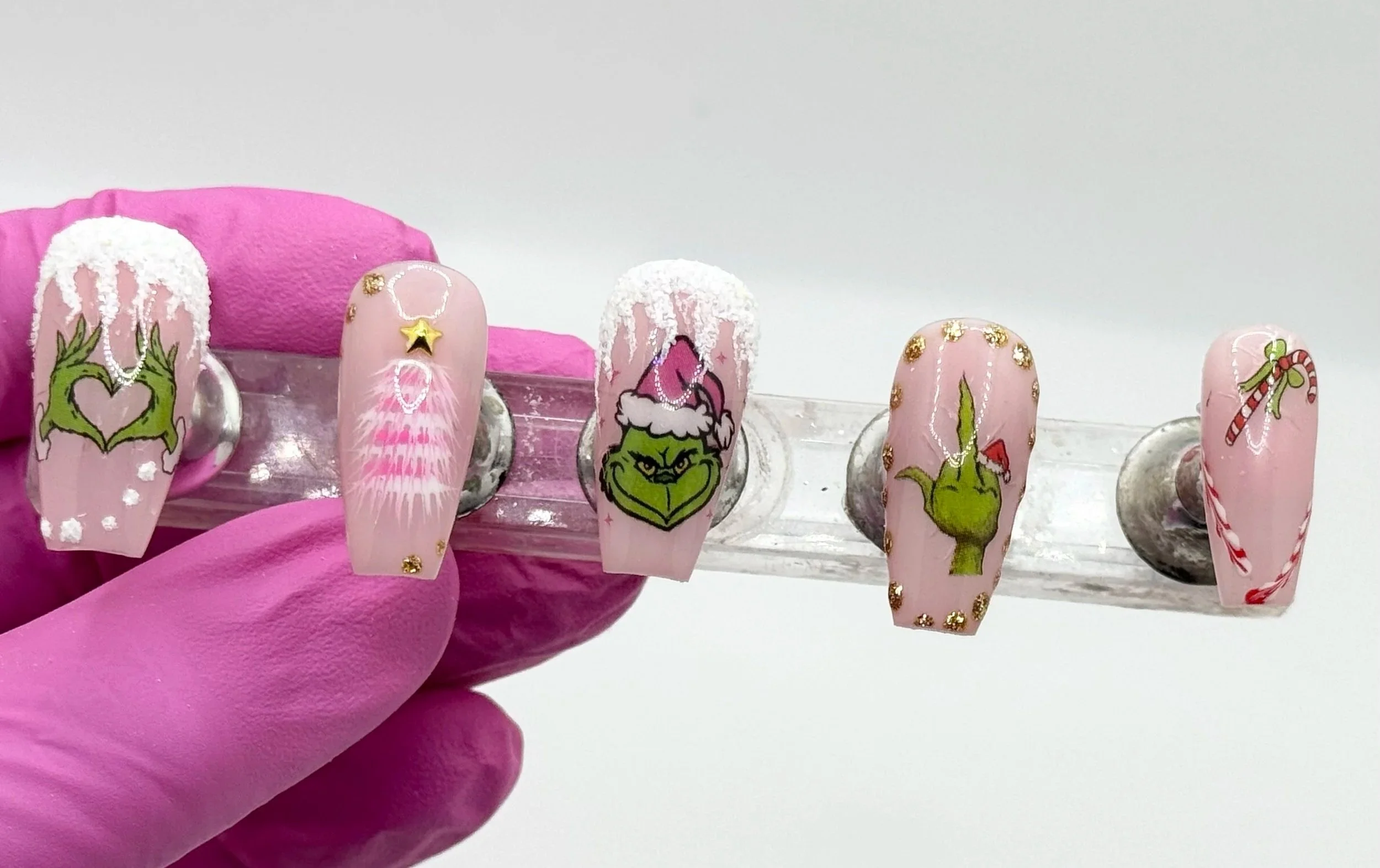 Pink Christmas press-on nails with glow-in-the-dark snow, gold accents, Grinch-inspired art, and pink tree designs