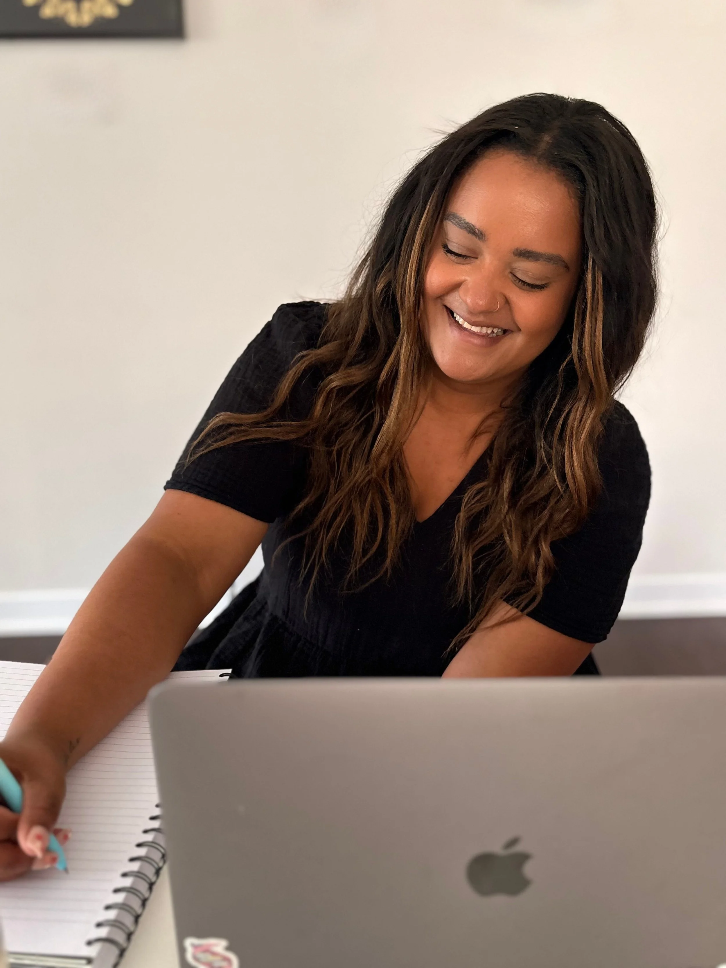 A woman with long, wavy dark hair and a bright smile sits at a desk with a silver Apple MacBook, a notebook, and a pen, looking at her laptop.