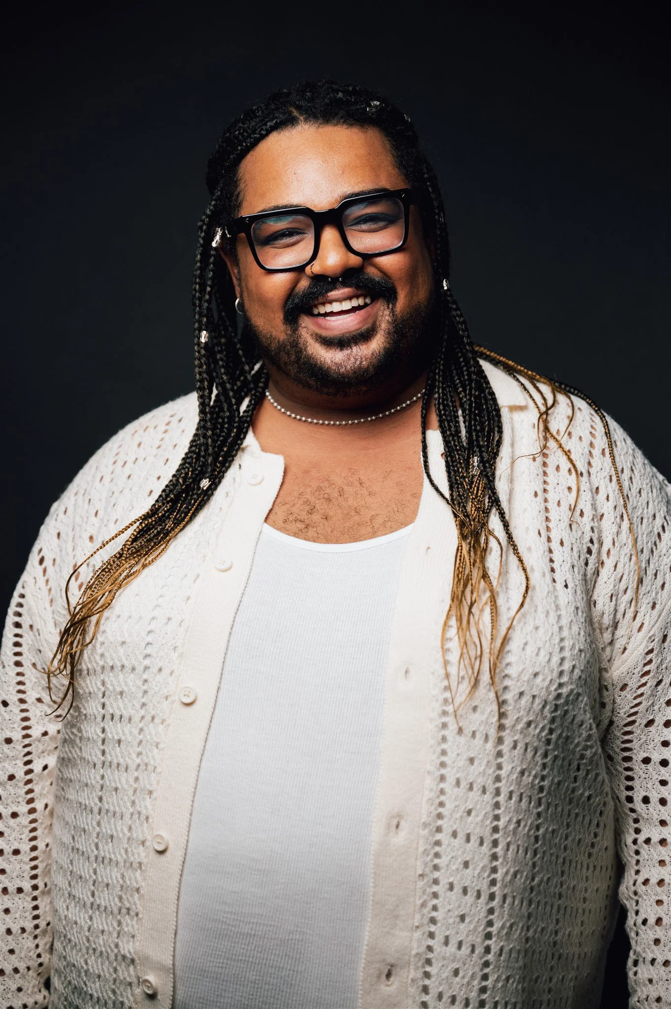 A smiling person with long braided hair, glasses, a nose ring, wearing a white tank top and a white crochet cardigan, standing against a dark background.