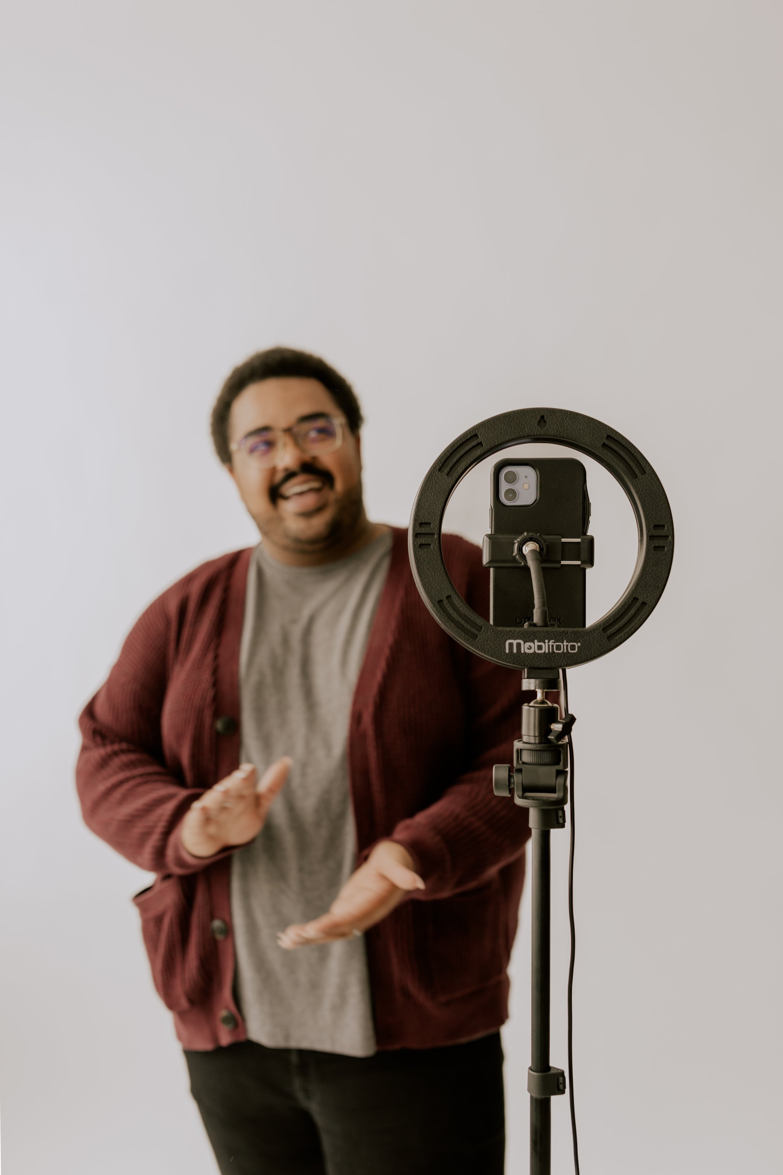 A man with glasses and a mustache, wearing a gray shirt and maroon cardigan, is talking while taking a selfie in front of a camera on a ring light stand, against a plain white background.