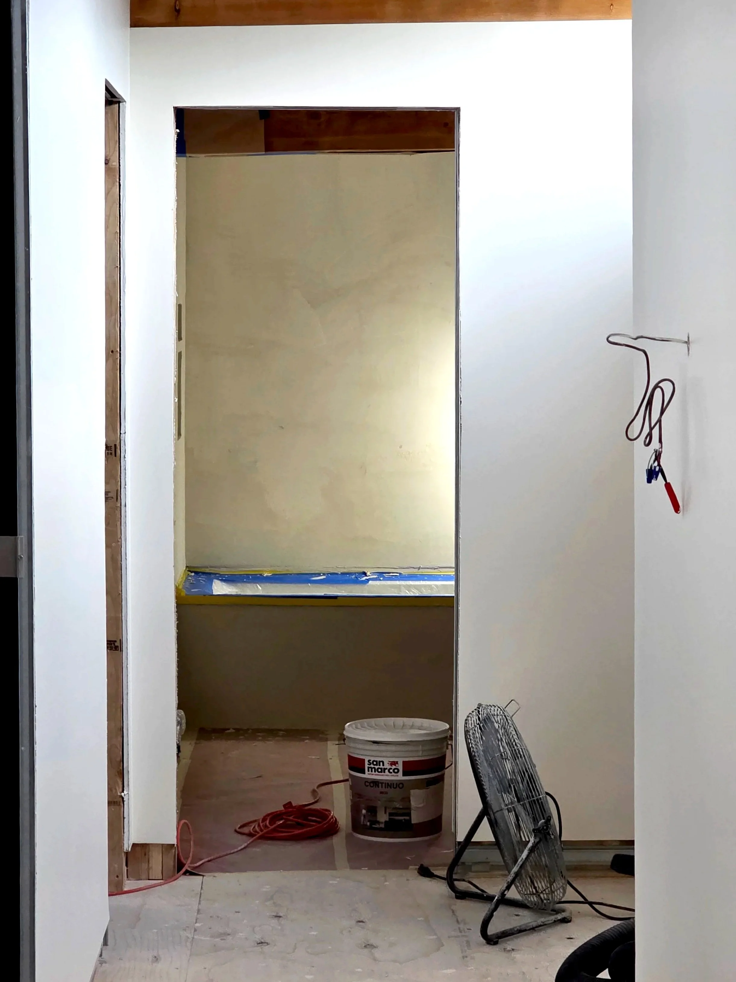 Interior of a room under construction with a white wall, plastic-covered opening, and construction tools including a fan, a bucket of microcement, and electrical cords.