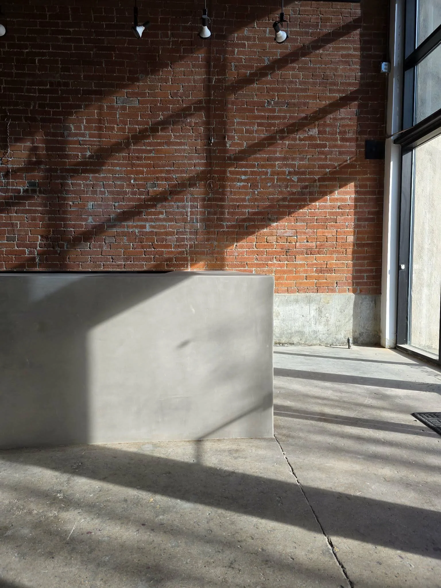 Sunlight creates shadows on a red brick wall, concrete floor, and a microcement counter inside a room with large windows.