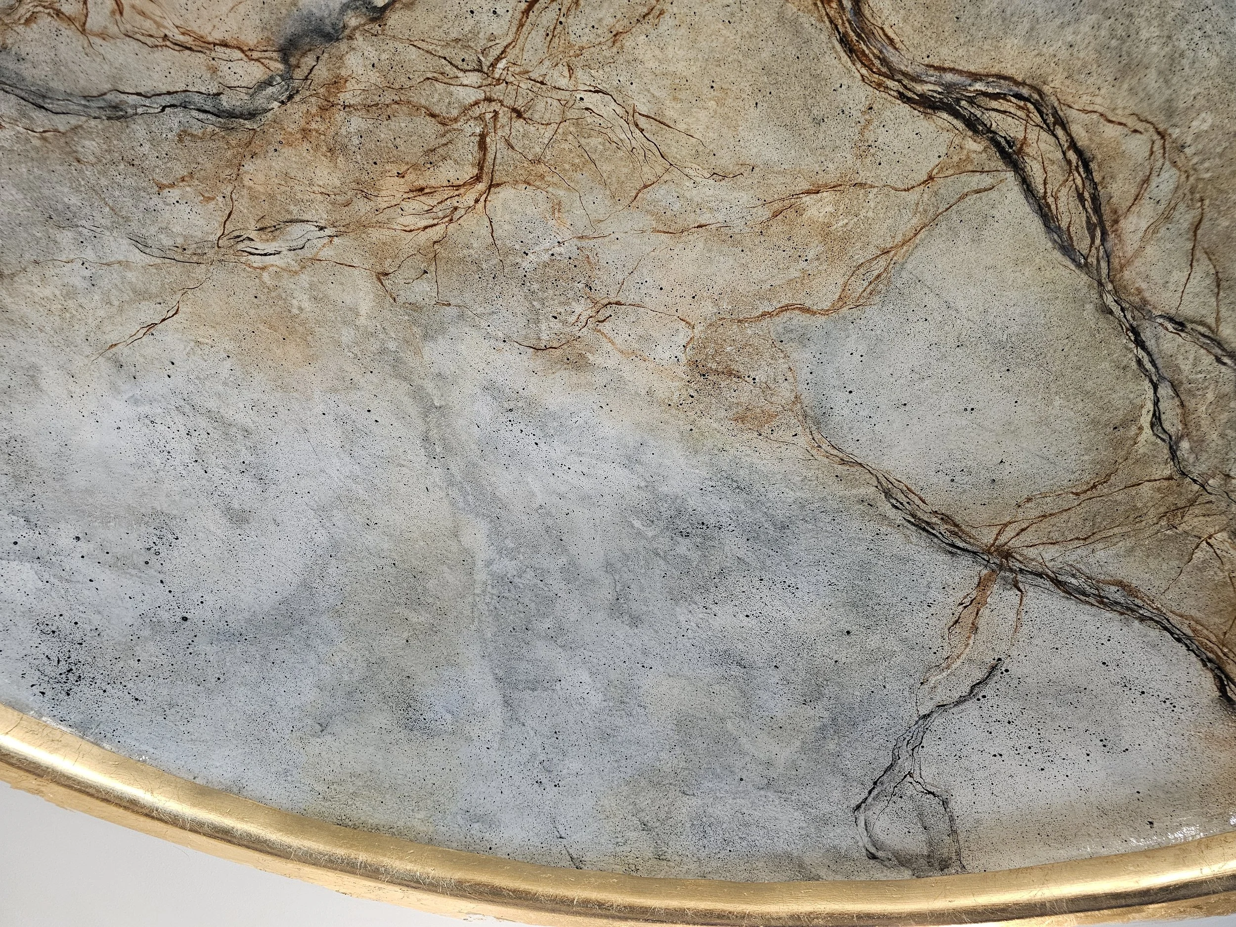 Close-up of a textured marble or stone surface with natural veining and speckles, containing light and dark brown veins and a thin gold border at the bottom edge.