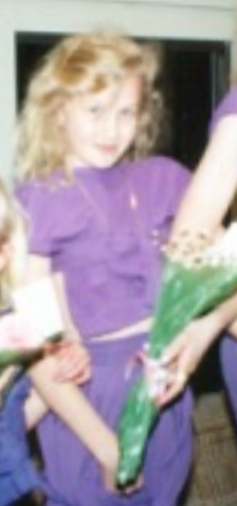 A young girl, Amie Drew Vreeken, MermaidSoundCave, with curly blonde hair wearing a purple shirt holding a bouquet of flowers after performing with her sister and cousin, a homemade production based on Disney's Aladdin. Costume design by Amie