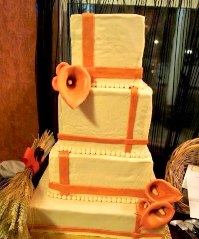 Three-tiered cake decorated with peach-colored fondant and large peach-colored calla lilies. The cake has orange fondant straps and white pearl-like decorations along the edges. It is displayed on a table with a woven basket and other items nearby.