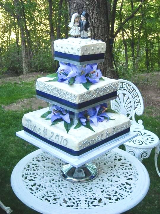 A three-tiered wedding cake with purple flowers, surrounded by a White outdoor setting with a white lattice table and chairs, topped with bride and groom figurines, set in a wooded area.