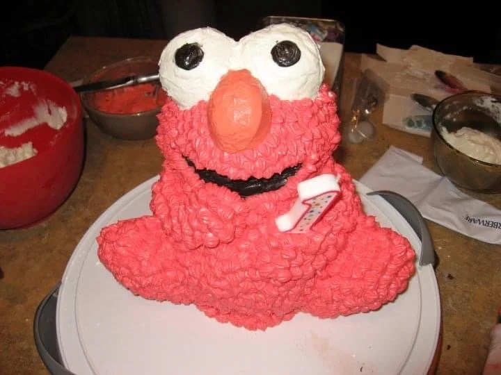 Cake shaped like Elmo from Sesame Street with red frosting, white eyes, an orange nose, and a black smile, decorated for a birthday celebration.