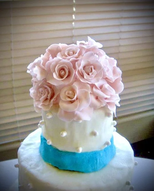 Two-tiered cake with white frosting, decorated with pink roses on top and blue fondant band around the middle, placed on a white surface in front of closed window blinds.