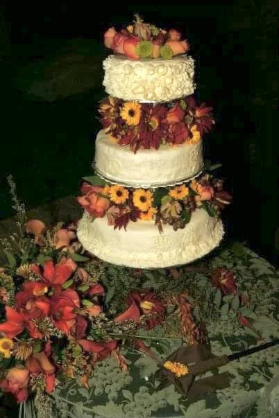 A four-tiered wedding cake decorated with assorted fresh flowers including sunflowers, roses, and other colorful blooms, set on a decorated table with scattered flowers and greenery.