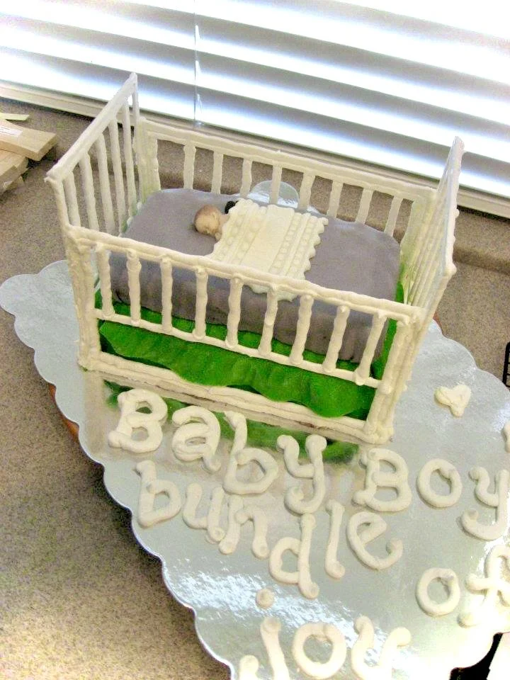 Cake designed to look like a baby's crib with a baby figurine inside, surrounded by green grass and a blanket, on a silver cake board with "Baby Boy birthday" written in white icing.