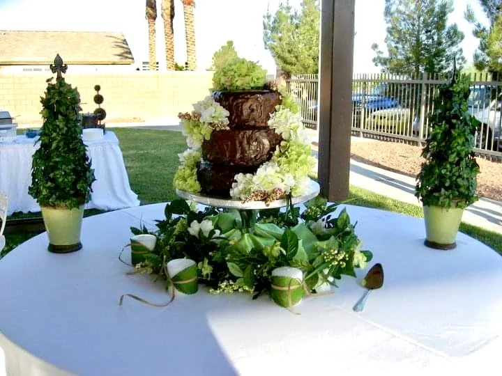 Chocolate wedding cake decorated with white and green flowers on a table with candle centerpieces and greenery, outdoors under a shaded area.