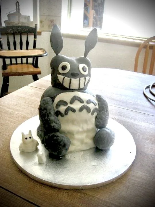 A cake shaped like Totoro with a large, smiling face, big eyes, and prominent teeth, sitting on a white base with a small Totoro figurine beside it, in a room with wooden chairs and a window in the background.