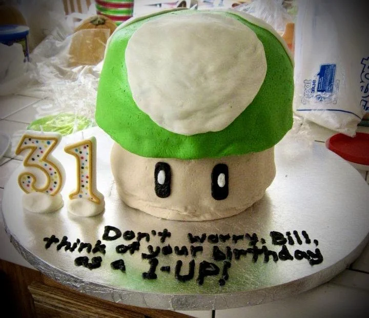 A cake designed to look like a Super Mario mushroom with a green cap and white spots, decorated for a 31st birthday, with candles shaped as the number 31 and a message that says "Don't worry, Bill, think of your birthday as a Y-UP!"