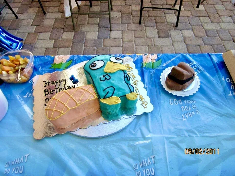 Celebration table with a birthday cake shaped like a blue bird, a cake with yellow crisscross icing, a chocolate dessert, and a bowl of fruit, on a blue tablecloth with chairs in the background.