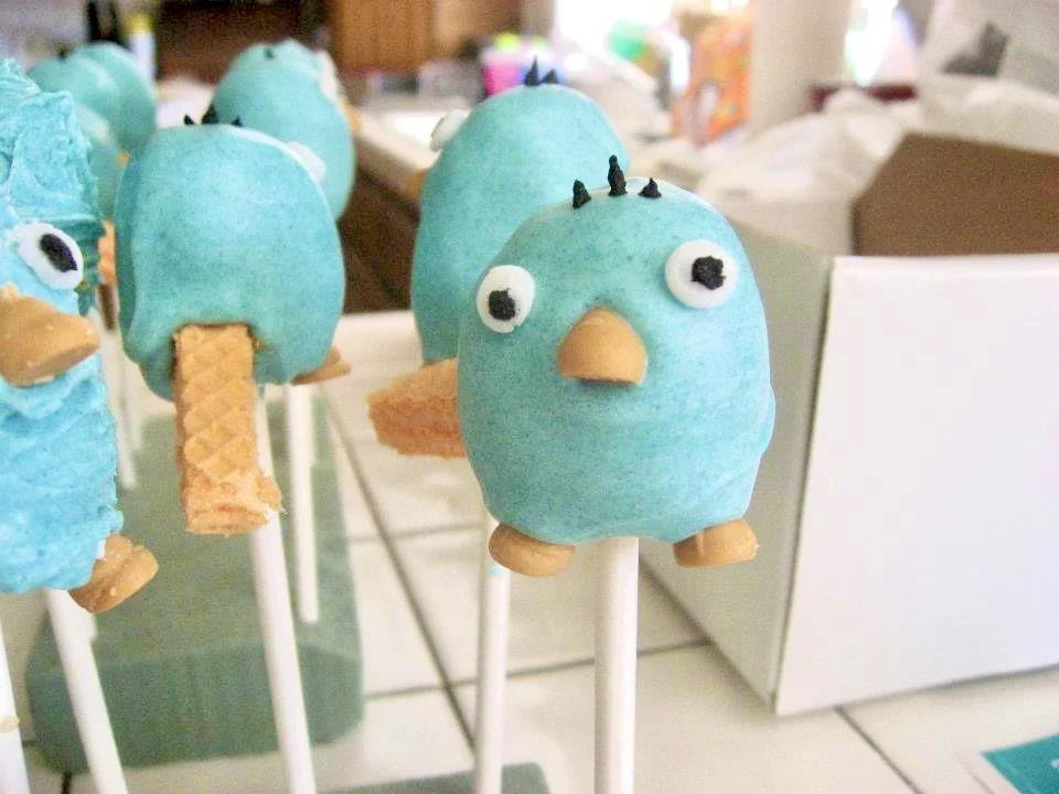 Cake pops decorated to look like cartoon birds, with blue coating, white eyes, orange beaks, and small black decorations on top, set on white sticks.