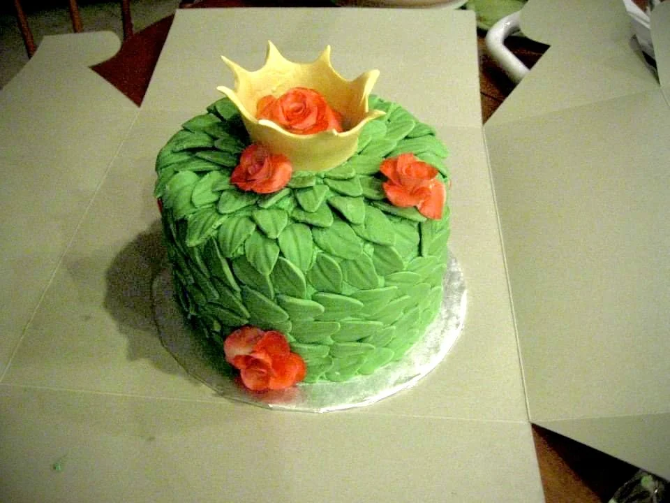 A round cake decorated with green leaves, orange flowers, and a yellow chalice with a red rose inside on top.