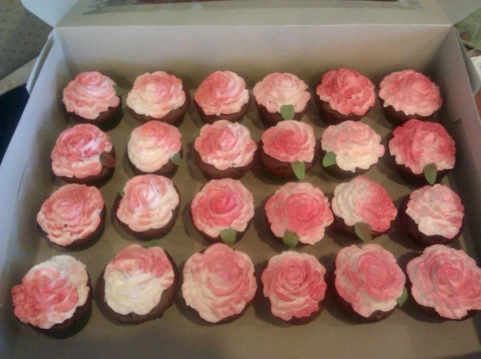 A box containing 25 cupcakes decorated with pink and white frosting to resemble roses, each with a small green fondant leaf.