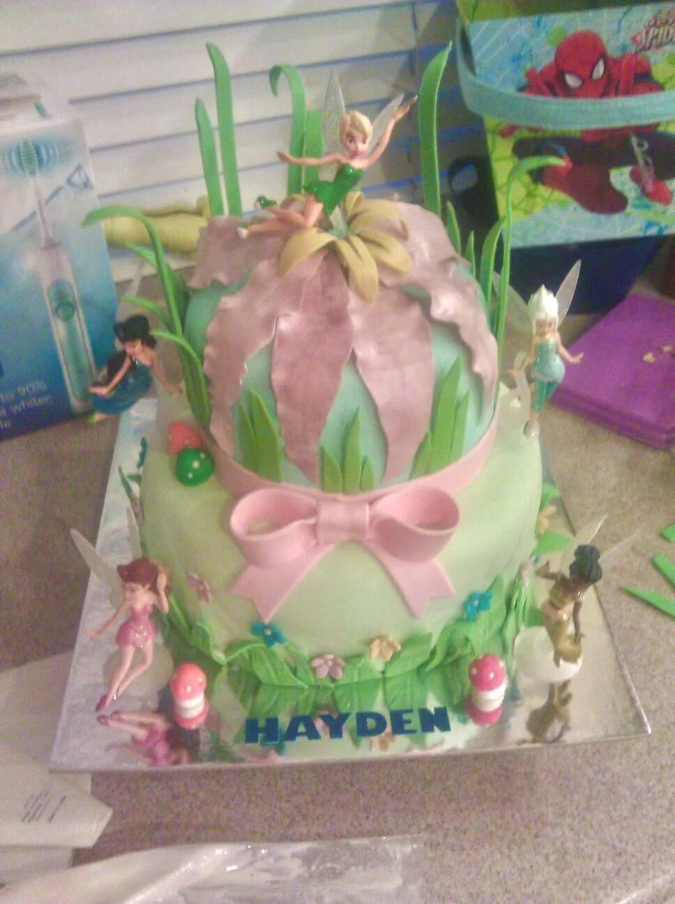 A two-tiered birthday cake decorated with a Tinker Bell theme, featuring Tinker Bell figurines, green grass-like decorations, pink bows, and the name Hayden on the front. There are additional fairy figurines around the cake, and a Spiderman-themed item in the background.