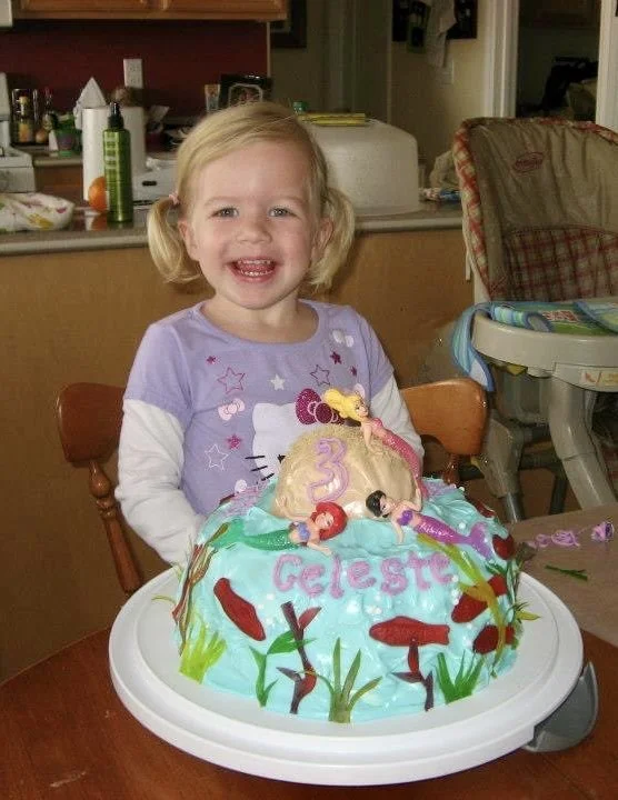 Young girl with blonde hair, smiling, standing behind a decorated Hello Kitty birthday cake on a table.
