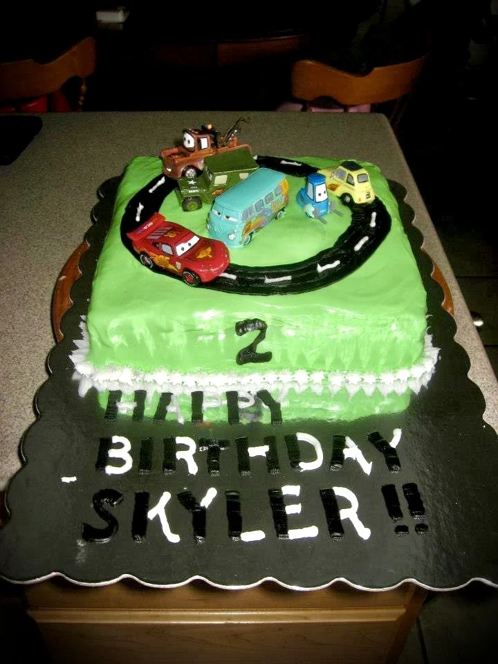 Green birthday cake with Cars characters on top, a black road with white lane markers, the number 2, and a message reading 'Happy Birthday Skyler!!' in black and white icing.