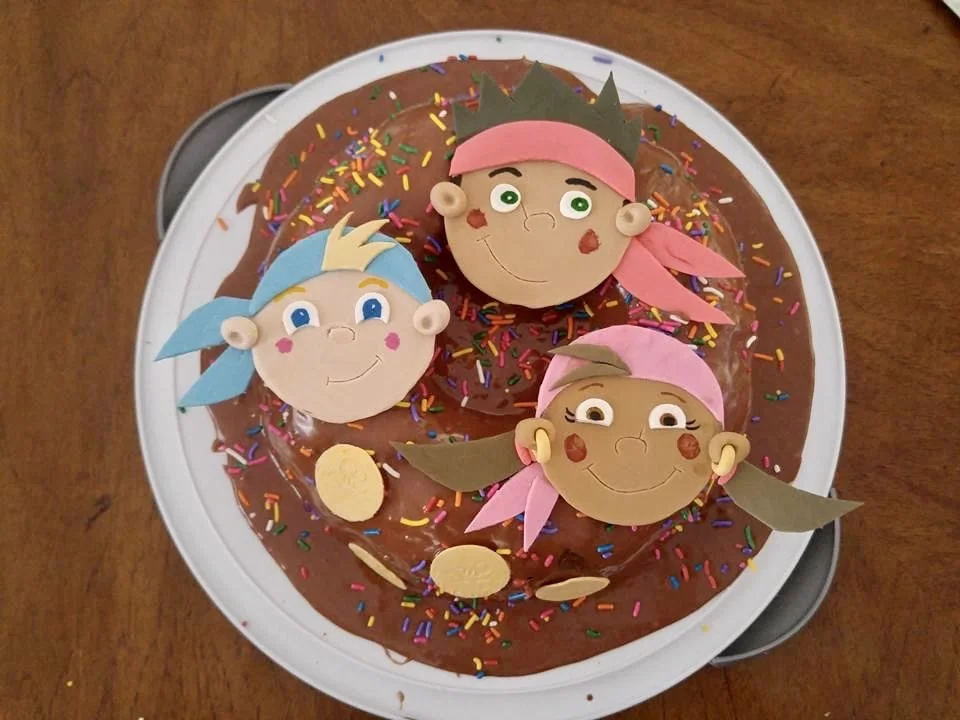 Chocolate cake topped with sprinkles and decorated with paper cutouts of three cartoon characters with different skin tones, hair accessories, and pink bandanas.