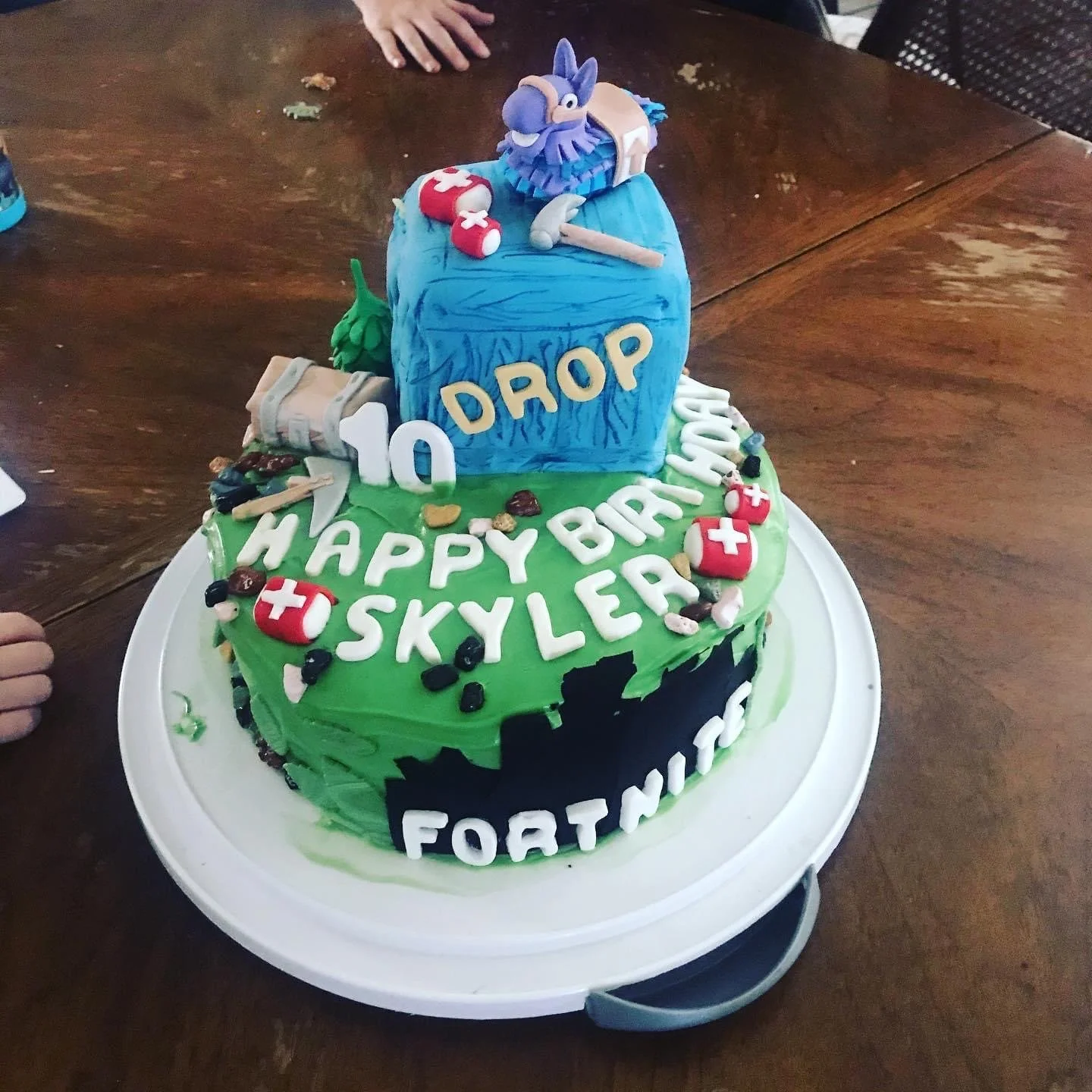 A two-tier birthday cake with Fortnite themes. The top tier is blue with a toy-like figure of a blue creature and gaming items. The bottom tier is green with edible trees, weapons, and small medical kits, decorated with white fondant letters spelling 'HAPPY BIRTHDAY SKYLER' and '10 DROP.' The cake also has a black fondant label with the word 'FORTNITE.'