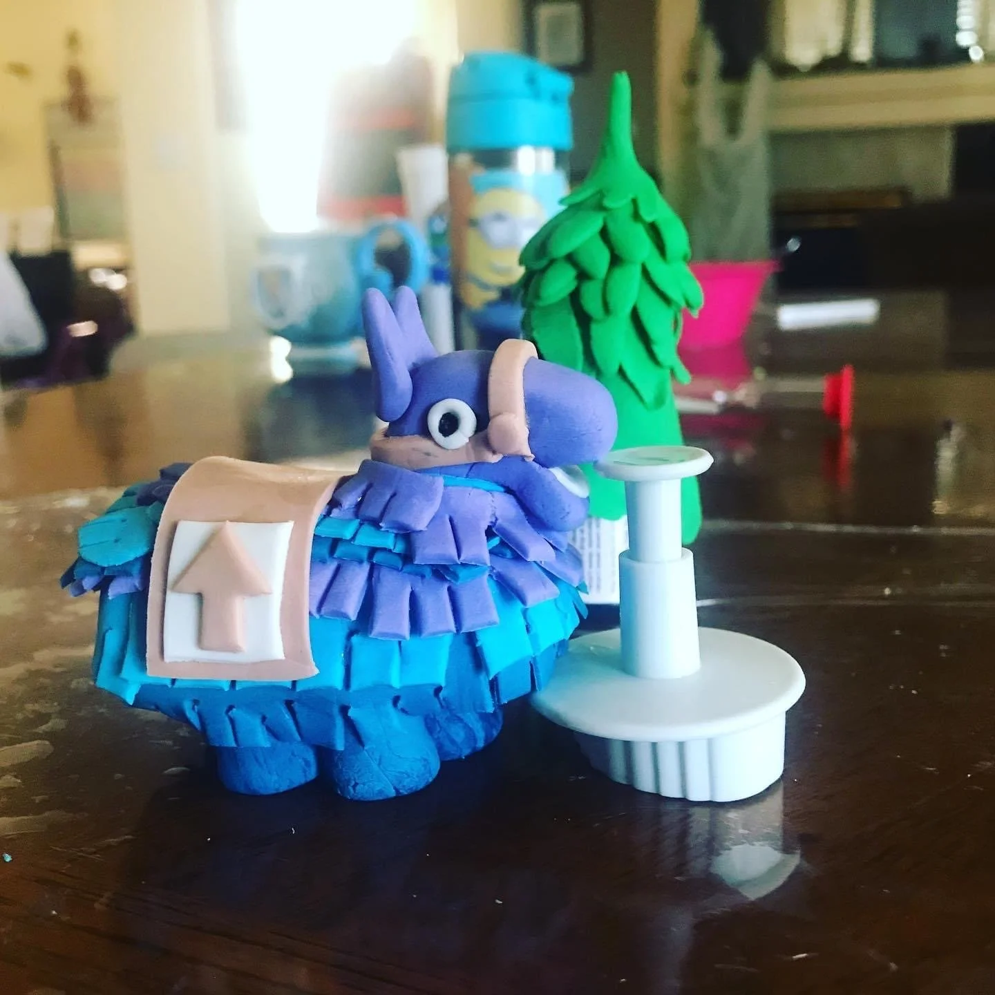 Clay art of a blue turtle with a shell decorated with purple and blue strips, wearing a tan backpack featuring a white upward arrow, with a green tree and a white lamp in the background.