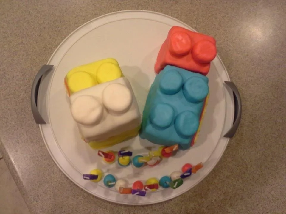 Colorful birthday cake shaped like LEGO bricks with a climbing bead toy around it, on a white tray.