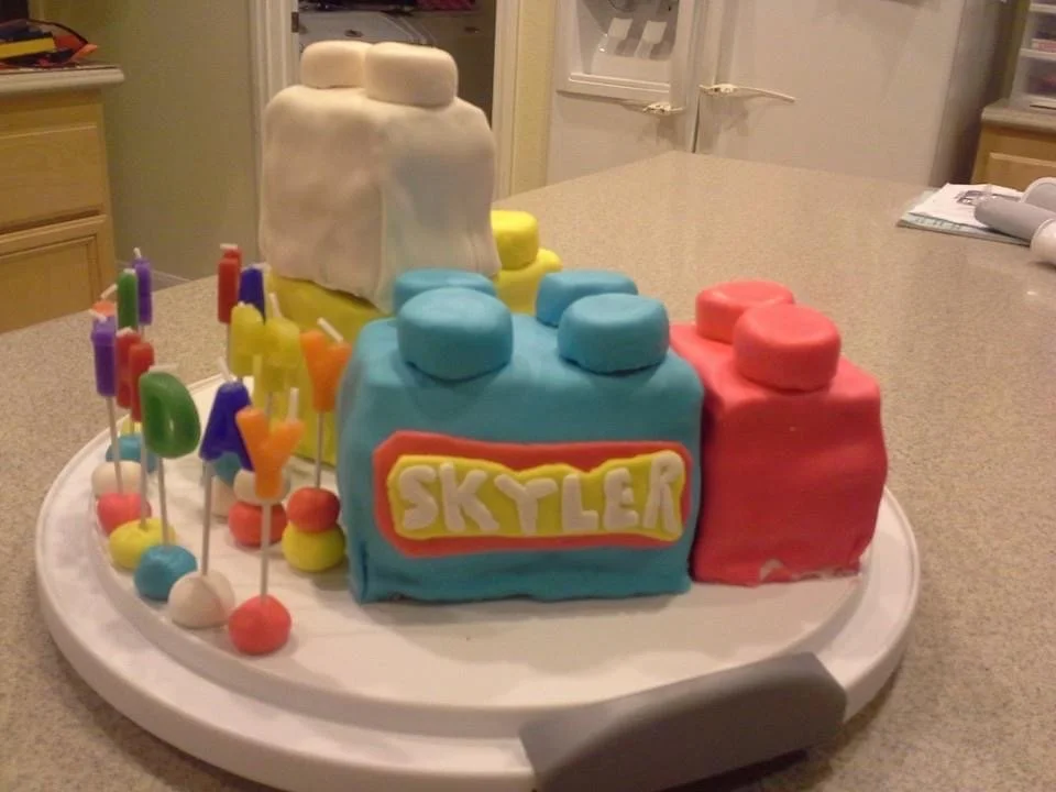 Colorful birthday cake shaped like a train with the name 'Skylear' on it, decorated with multiple pastel-colored fondant blocks and candles on a white tray.