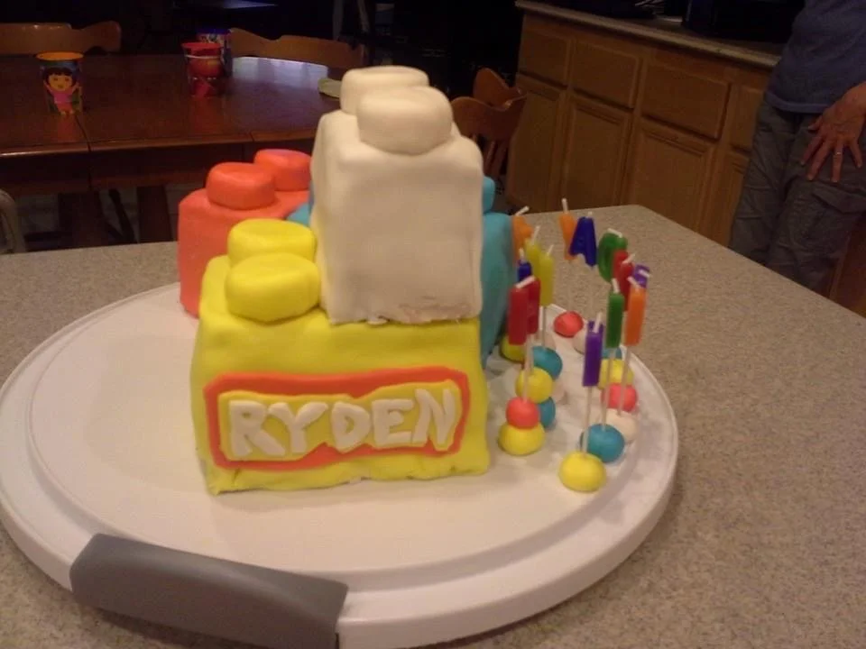 Colorful birthday cake with the name 'Ryden' on the front, surrounded by decorated candies and candles, on a white cake tray.