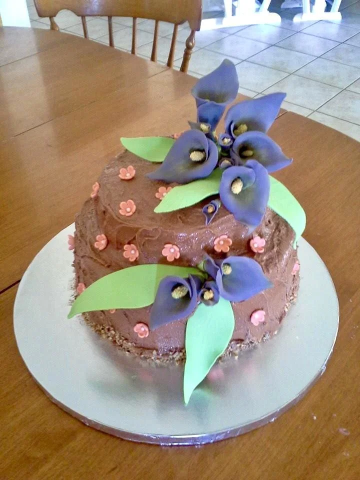 Chocolate birthday cake decorated with purple flowers and green leaves on a wooden table.