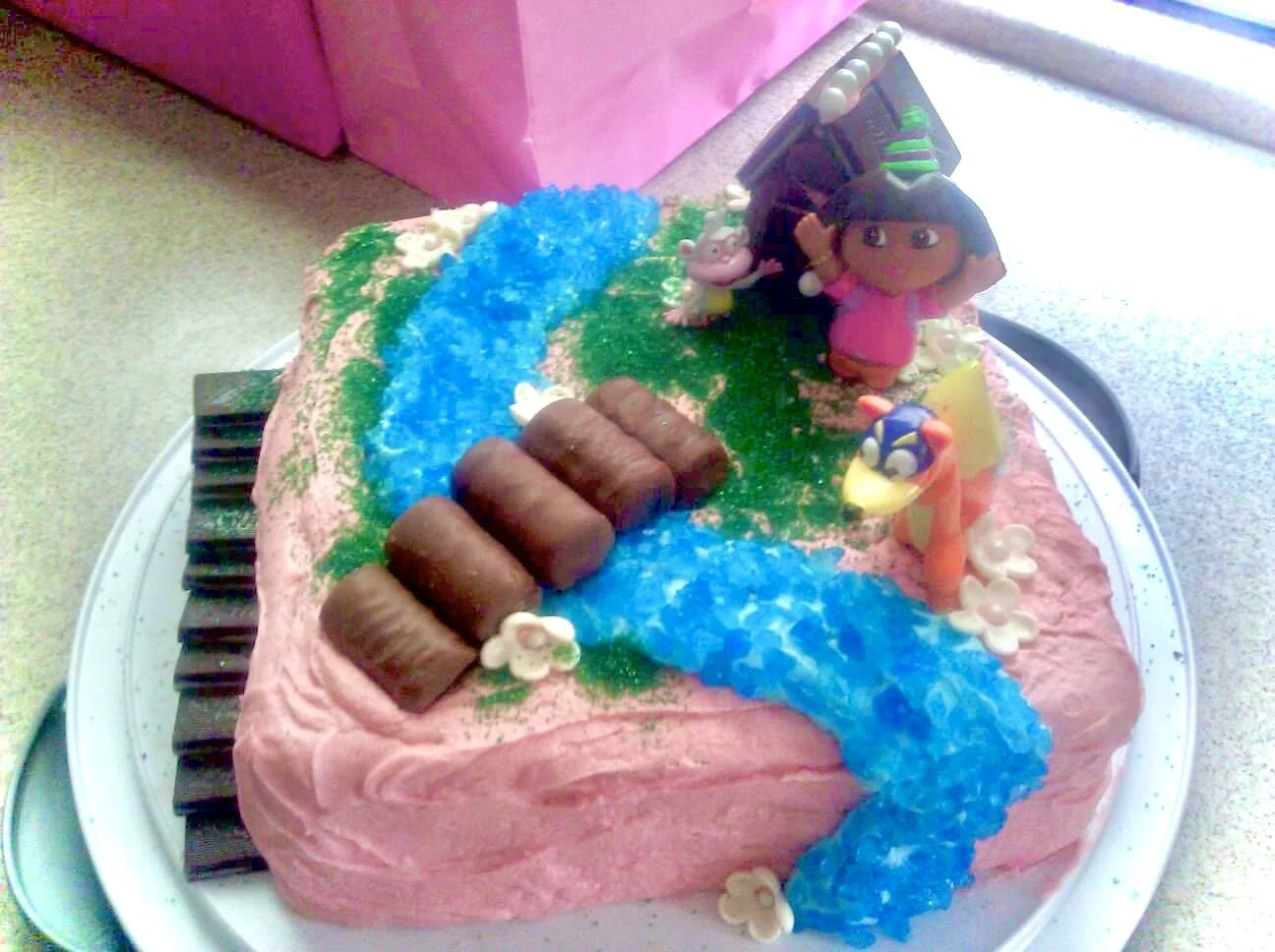 A pink foam cake decorated with a blue candy river and green grassy area, featuring fondant figures of Dora the Explorer, her monkey friend Boots, and a fox.