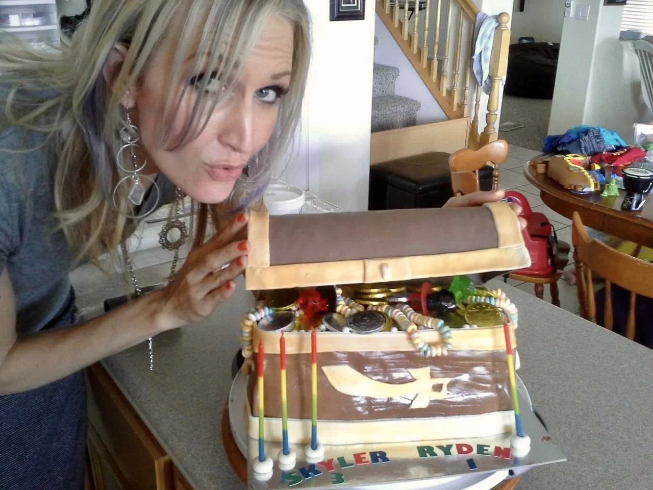 A woman with blonde hair and hoop earrings revealing a birthday cake designed as a treasure chest filled with jewelry.